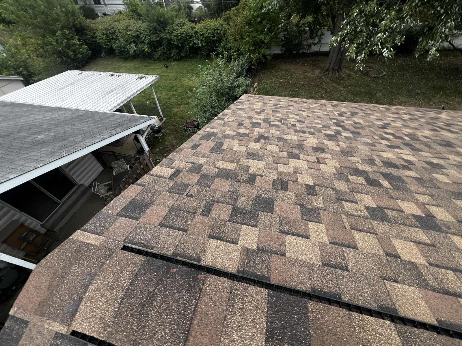 Wide overhead view of a newly laid brown, tan and charcoal architectural asphalt shingle roof plane filling the right of the frame, with a lower grey shingled porch roof and a white corrugated metal carport canopy on slim white posts at the left, and a back yard below with a kettle barbecue, patio chairs, hedges and a large tree