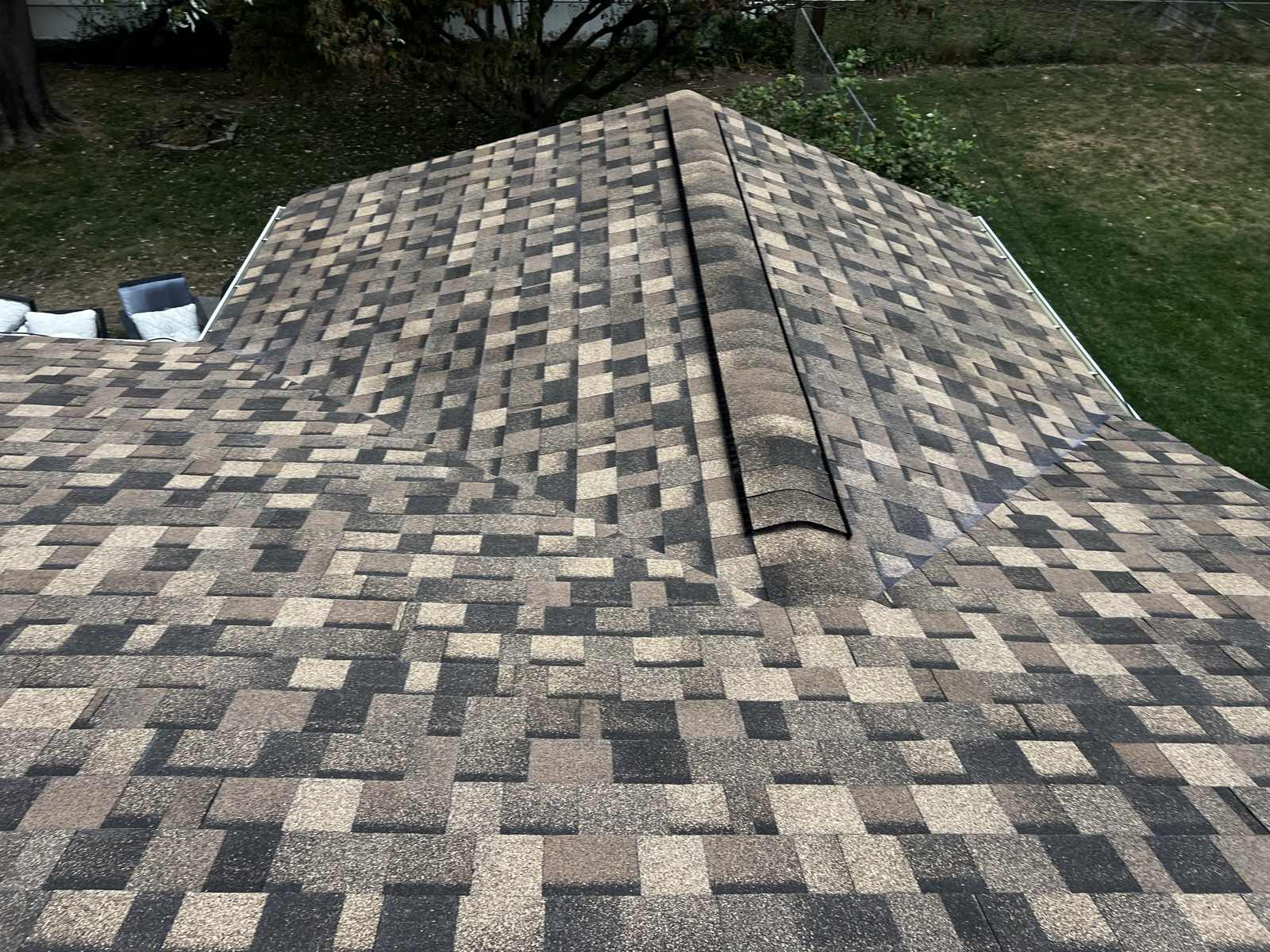 Overhead view of the hip of a newly laid brown, tan and charcoal multi-tone architectural asphalt shingle roof, capped along the hip with ridge cap shingles and carrying a dark ridge vent strip, with the two roof planes falling away either side, and below a mown lawn, a chain-link fence, a bare-branched tree and outdoor cushioned furniture