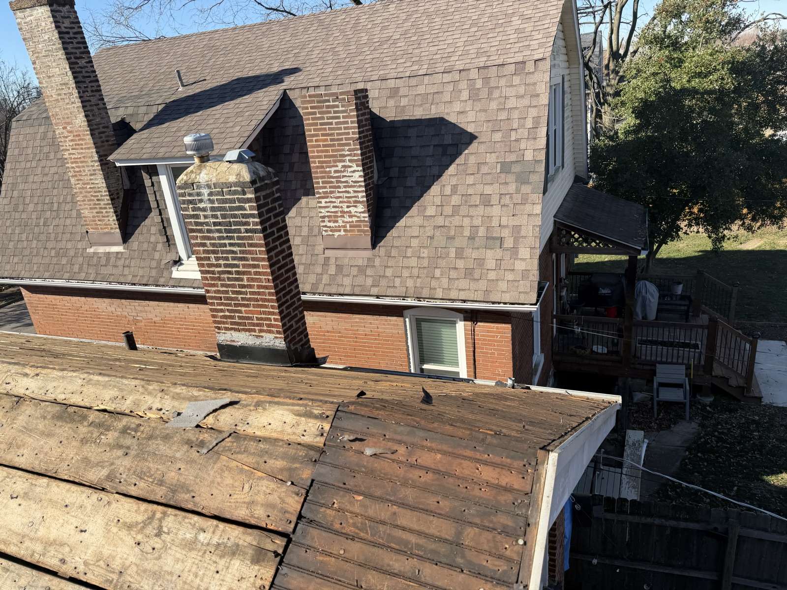 A shallow pitched lower roof plane in the foreground stripped back to dark weathered plank decking with scraps of old felt still attached, running out to a white gutter, while behind it the main red brick house still carries its full weathered brown asphalt shingle roof, with two brick chimneys showing spalled and patched mortar, a dormer with a metal turbine vent, and a covered wooden rear deck at the right