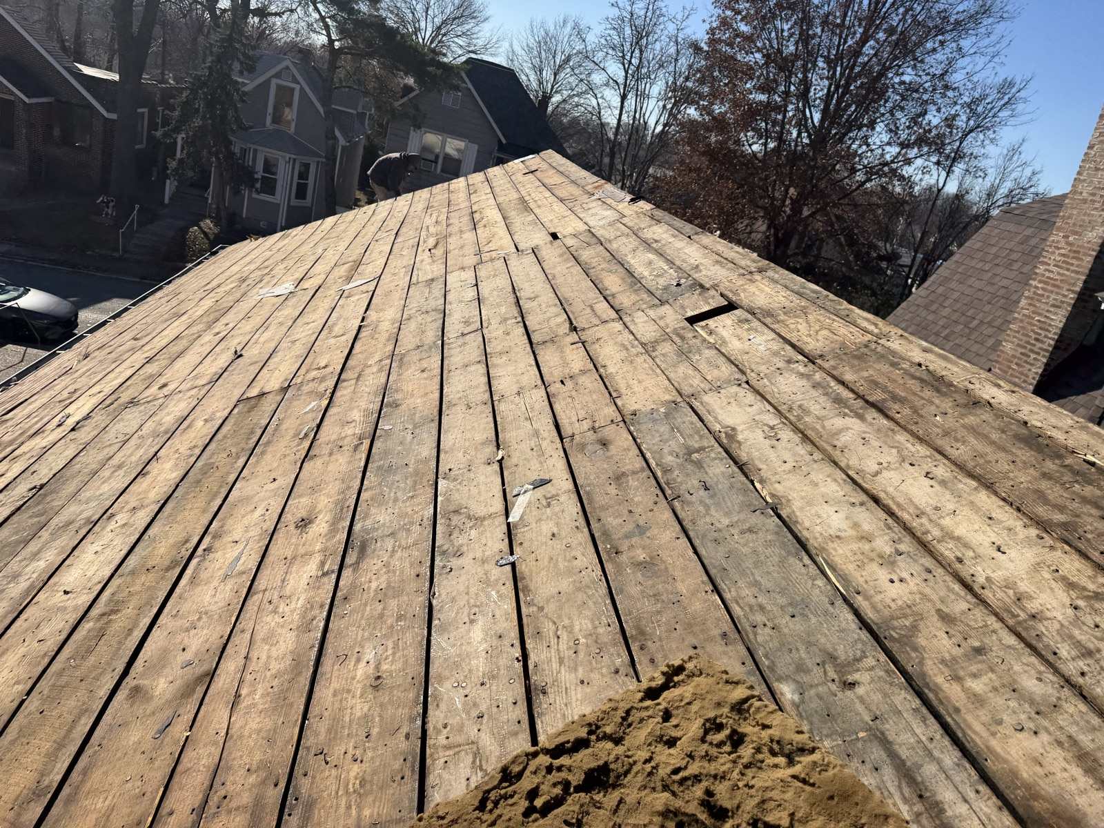 A large roof plane stripped back to bare wooden plank decking filling most of the frame, the boards weathered and studded with old nails with visible gaps between them, scraps of felt caught in the joints and a pile of loose granules and debris at the lower right, with a figure bent over at ground level beyond the far ridge beside a neighbouring grey house, a grey-blue two-storey house with a bay window and a brick house beyond under a clear winter sky