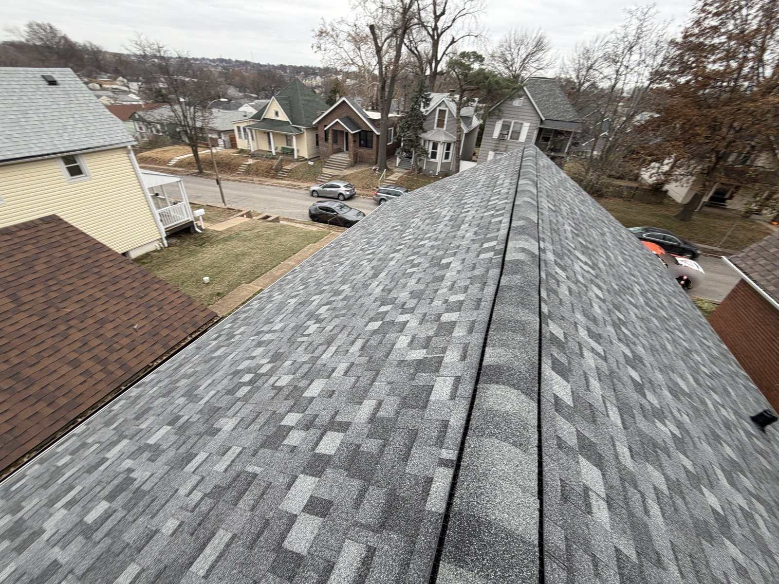 Newly laid grey and charcoal architectural asphalt shingle roof photographed along its ridge, with a dark ridge vent strip running down the centre, looking out over a St. Louis street of close-set older houses including a yellow two-storey, a green house with a turret and a brick bungalow, with cars at the kerb and wooded hills on the horizon