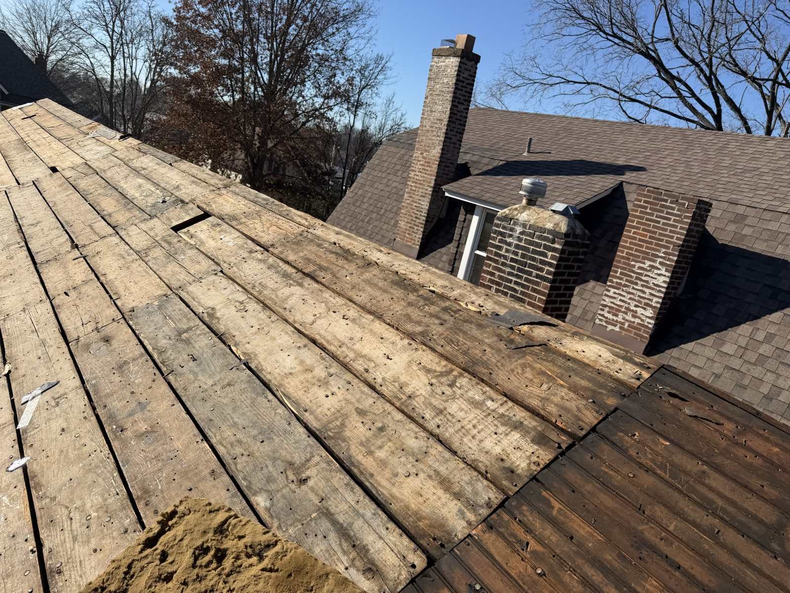 Stripped wooden plank decking running up to the left of the frame with the boards sitting at visibly uneven heights, one board displaced and a gap open in the run, old nails throughout, and beyond it the main house still carrying its weathered brown asphalt shingles with two brick chimneys showing spalling and missing mortar and a small dormer with a metal turbine vent