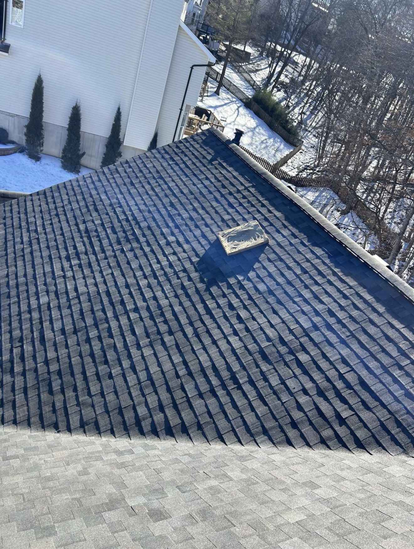 Portrait view of a roof mid installation with courses of dark charcoal architectural asphalt shingles laid down the upper plane, casting hard shadow lines, meeting a lighter grey shingled surface across the lower part of the frame, an offcut lying loose on the slope, and beyond the roof edge a cream vinyl sided house, a picket fence, a raised deck and snow lying on the ground under bare winter trees